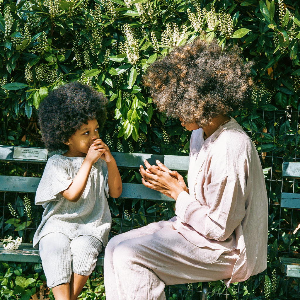 A mother and her young child sit facing each other on a sunlit garden bench, deep in conversation.